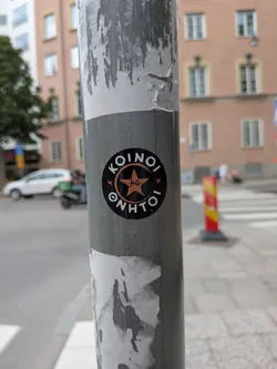 Street sticker A round black sticker affixed to a metal pole. It features the white Greek text 'KOINOI ΘΝΗΤΟΙ' wrapping around a central copper-colored star emblem containing the initials 'KΘ'.