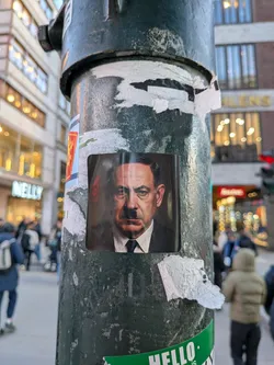 The sticker is a rectangular portrait of a man with dark, slicked-back hair and a distinctive 'toothbrush' mustache. He wears a dark suit, a white collared shirt, and a dark tie, looking directly forward with a stern expression. The background is dark, possibly a deep red or brown, giving the image a somber tone. The sticker appears to be a printed photograph or realistic illustration, adhered to a textured, dark green metal pole.
