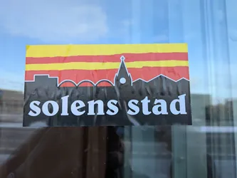 Street sticker The sticker is rectangular, featuring a horizontal striped pattern at the top with alternating yellow and red bands. Below this, a black silhouette depicts a city skyline, prominently featuring a tall church spire with a cross at its peak and a circular element (possibly a clock or window). Other buildings with varying rooflines are visible in the silhouette, outlined in white. The bottom section of the sticker is black, with the words "solens stad" written in large, bold white sans-serif letters. The sticker appears to be applied to a glass surface, showing some minor wrinkles and reflections.