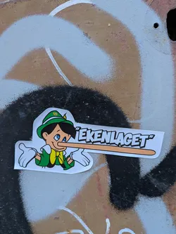 Street sticker The sticker features a cartoon character resembling Pinocchio. He is depicted wearing a green hat with a yellow band and a white feather, a green jacket, a yellow scarf, and white gloves. His signature long nose is prominently extended, suggesting he is telling a lie. The character's eyes are blue and he has a slight smile. The sticker has a white border and is placed on a background with graffiti.