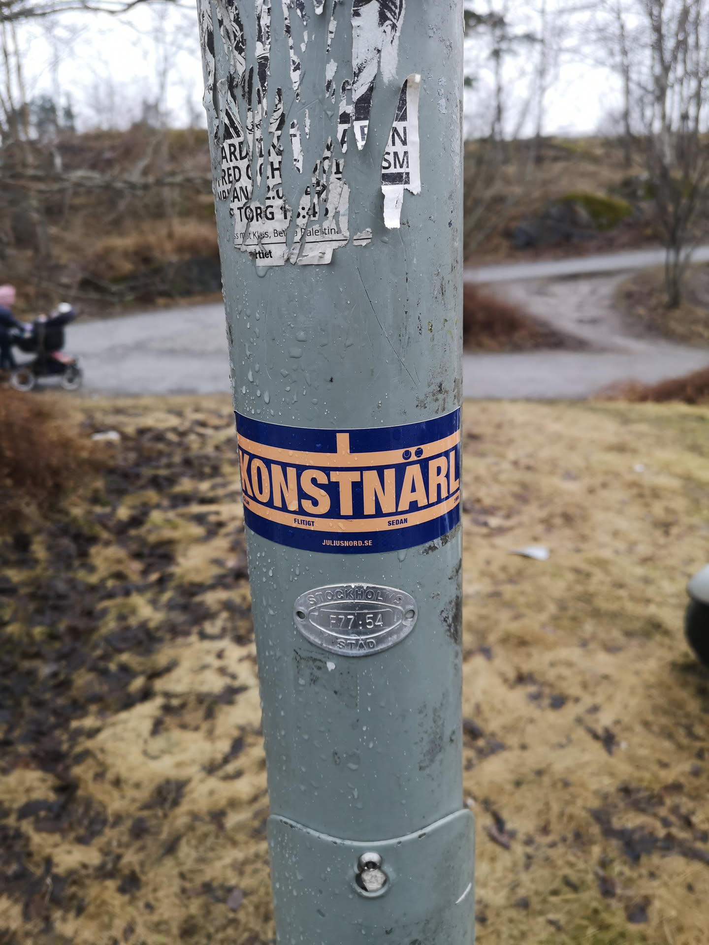 The sticker is a horizontal band, predominantly dark blue, with a central orange stripe. Within this orange stripe, the word 'KONSTNÄRL' is prominently displayed in bold, orange, sans-serif capital letters. Above the 'Ä' are two small dots. Thin orange lines border the top and bottom of the central orange stripe. Below the main text, in smaller white or light-colored font, 'FLITIGT' is on the left and 'SEDAN' on the right. Centered beneath these words is 'JULIUSNORD.SE'. The sticker is applied to a grey pole and shows signs of being outdoors, with visible water droplets.