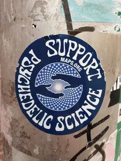 Street sticker A round sticker with a dark blue background and white text. The text says "Support Psychedelic Science" in a circular arrangement. Inside the circle is a design of two hands reaching towards a bright light, surrounded by a dotted pattern. The text "MAPS.ORG" is visible in smaller font inside the main text.