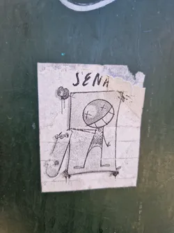 Street sticker A hand-drawn sticker featuring a simple character design. The character seems to be riding a skateboard. The style is minimalistic, with basic shapes and lines. The sticker has some tears and creases, suggesting it might have been placed outdoors for some time.  The name 'J'ENA' is written at the top.