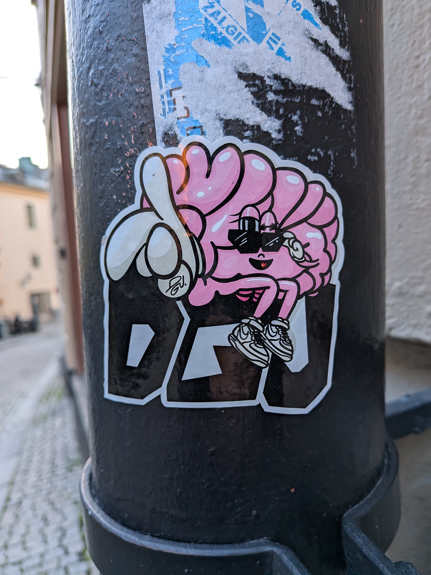 Street sticker The sticker features a stylized, anthropomorphic brain character, predominantly pink with lighter pink highlights and a thick black outline. The brain has a friendly, smiling face with a small red mouth and is wearing black rectangular sunglasses. It has cartoonish arms and legs; one hand is giving a thumbs-up gesture, and its feet are clad in black and white striped sneakers (resembling Nike Dunks or similar). The character is positioned above the bold, block letters "DEU" which are black with a white outline. A small, stylized signature "GU." is visible on the thumb of the hand giving the thumbs-up. The sticker has a clean white border around its entire perimeter and is affixed to a textured black pole.