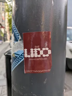 A rectangular sticker with a dark reddish-brown (maroon) background. The text is rendered in white. At the top, 'BAR' is written in a standard sans-serif font. Below it, 'LIDO' is prominently displayed in a unique, highly stylized rounded font where each letter is composed of interconnected circular or oval shapes. Further down, 'www.barlido.se' is written in a smaller, standard sans-serif font, followed by 'DROTTNINGGATAN 82' at the bottom, also in a standard sans-serif font. The sticker has a slightly glossy finish and shows minor wear along its edges.