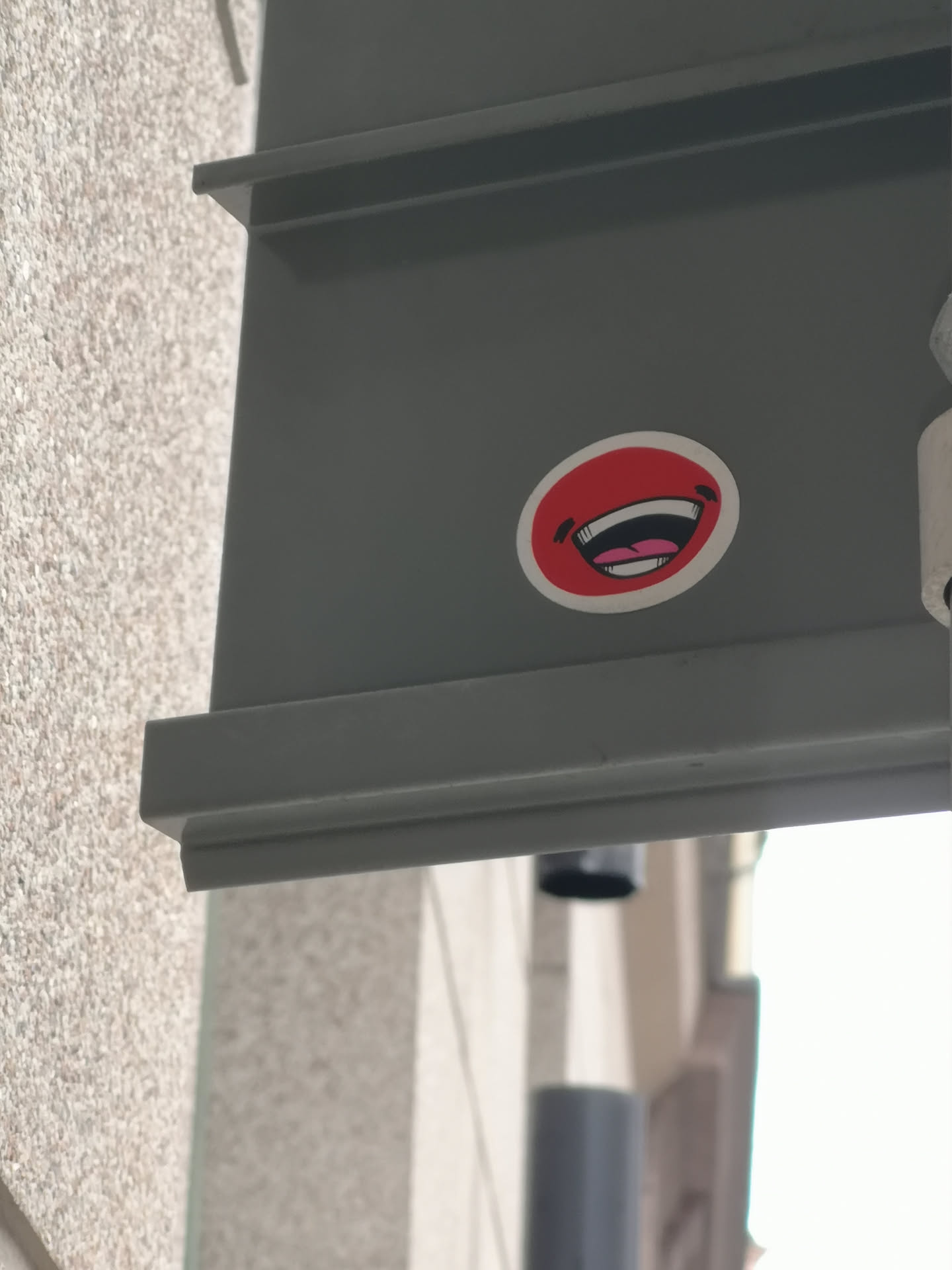 The sticker is an oval shape with a vibrant red background, encircled by a clean white border. Dominating its center is a stylized, wide-open mouth, depicted in a clear laughing expression. The mouth cavity is rendered in black, contrasting with bright white teeth and a visible pink tongue. Small, curved black lines are positioned on either side of the mouth, suggesting crinkled eyes or cheeks, which amplify the joyful and humorous appearance.