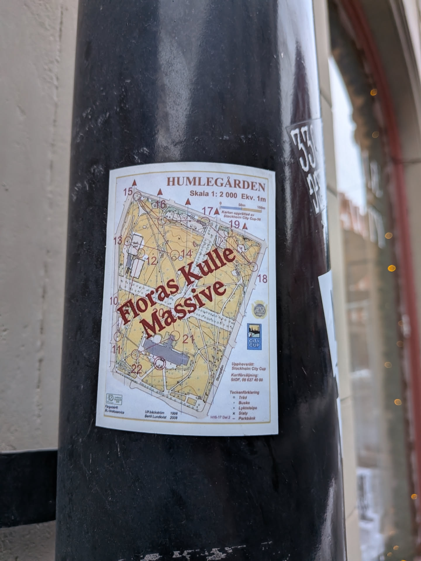 A rectangular sticker featuring a detailed orienteering map of Humlegården park. The map has a white background with yellow-beige areas for open ground, green for vegetation, and grey for buildings. Brown contour lines and black paths are visible, along with numbered control points (10-22) and red triangles indicating features. The prominent text 'Floras Kulle Massive' is written diagonally across the map in large, bold red letters. The sticker also includes a scale bar, a legend for map symbols (trees, bushes, lampposts, statues, park benches), and information about its origin, including 'Stockholm City Cup' and names 'Ulf Bäckström 1998' and 'Bertil Lundqvist 2008'. Small logos for 'City Cup' and another organization are present. The sticker is affixed to a dark, textured pole and shows slight curling at the edges, indicating it's a physical print.