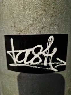 A black sticker with white graffiti-style writing. The word "tast" is written with an arrow pointing to the right.