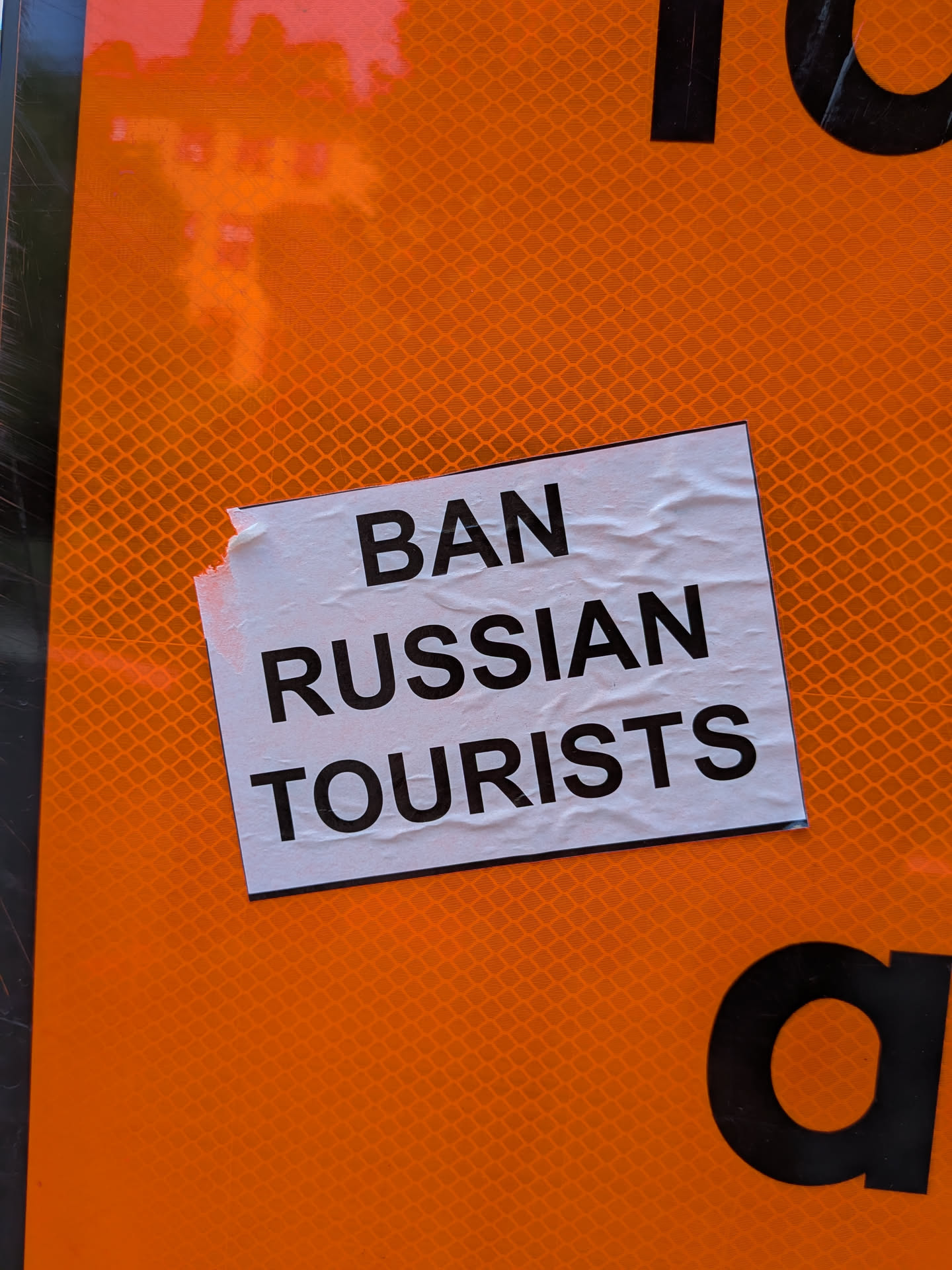 A rectangular white sticker with bold black text reading 'BAN RUSSIAN TOURISTS' in all caps. It is pasted on a reflective orange sign and shows slight wear on the left edge.