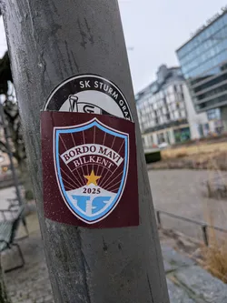 Street sticker The sticker is a shield-shaped crest, primarily in a deep maroon (bordo) color with light blue and white accents. It features a light blue border around the shield. Inside the shield, the words "BORDO MAVI" are arched at the top, with "BILKENT" directly below them, all in white text. Below the text, a golden-yellow five-pointed star is centered, from which white rays emanate downwards against the maroon background. At the very bottom of the shield, on a light blue band, the year "2025" is written in white, and below that, a stylized 'TS' logo in light blue is set against a white background. The sticker has a slightly glossy finish and shows visible water droplets on its surface, indicating it's exposed to the elements.