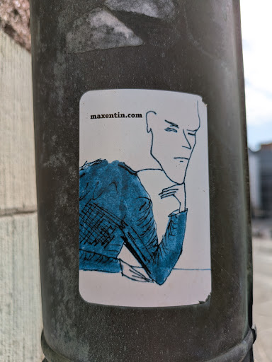 Street sticker The sticker depicts a minimalist line drawing of a man resting his chin on his hand, dressed in a blue jacket. The sticker is stuck to a grey pole. The website maxentin.com is written in the top left corner.