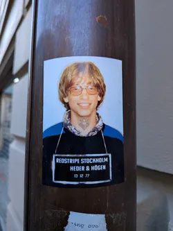 Street sticker The sticker is a rectangular print with slightly rounded corners, featuring a headshot of a young man strongly resembling a youthful Bill Gates. He has shaggy light brown hair, wears large amber-tinted glasses, and is smiling broadly. He is dressed in a dark blue and black collared shirt with a patterned inner collar. A distinctive, intricate black tattoo, possibly tribal or geometric in style, is visible on his neck just below his chin. The background of the portrait is a light blue. Below the portrait, there is a black rectangular bar with white text, mimicking a mugshot identification board. The sticker is affixed to a dark brown, possibly metallic or wooden, pole.