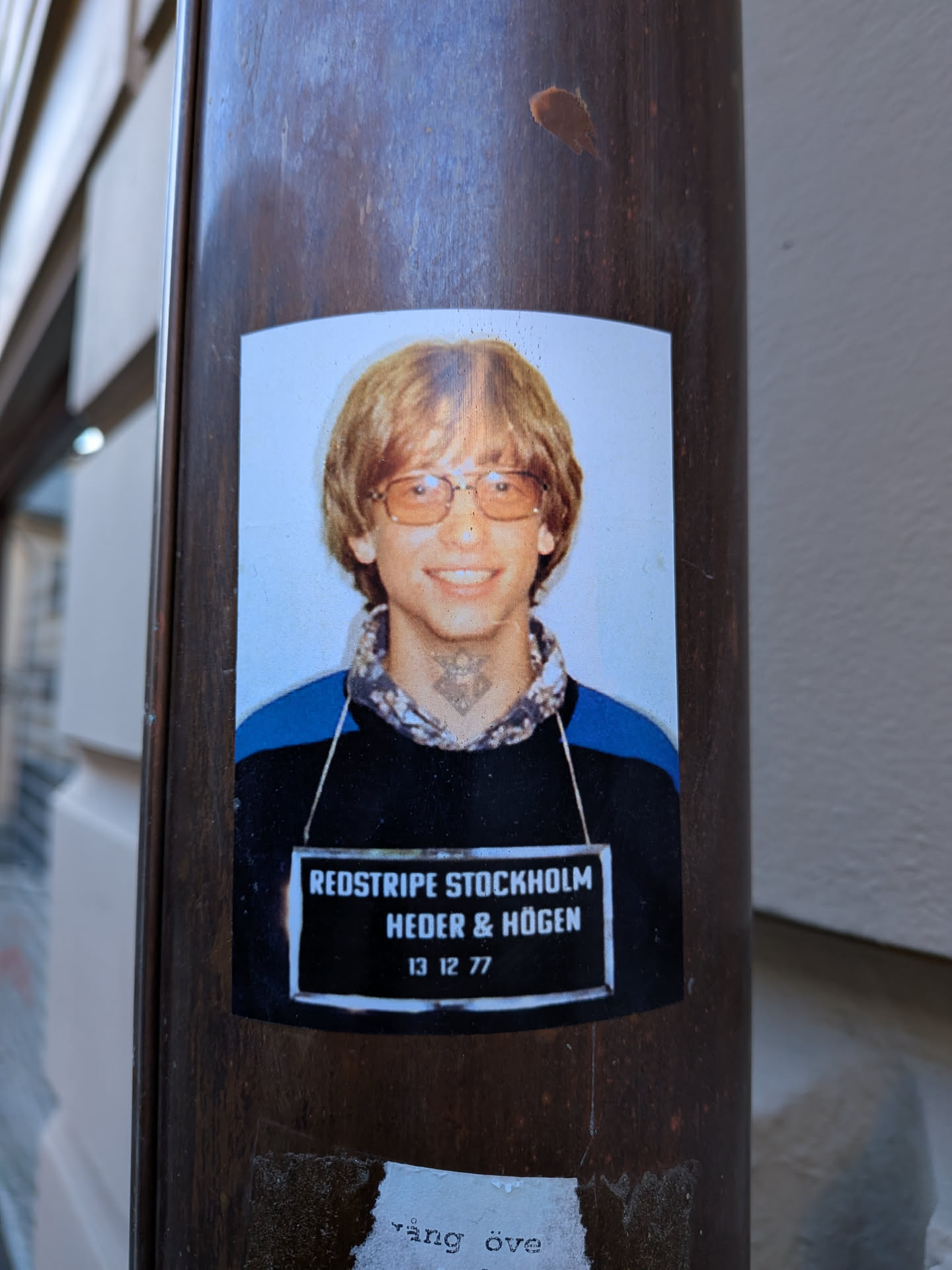 Street sticker The sticker is a rectangular print with slightly rounded corners, featuring a headshot of a young man strongly resembling a youthful Bill Gates. He has shaggy light brown hair, wears large amber-tinted glasses, and is smiling broadly. He is dressed in a dark blue and black collared shirt with a patterned inner collar. A distinctive, intricate black tattoo, possibly tribal or geometric in style, is visible on his neck just below his chin. The background of the portrait is a light blue. Below the portrait, there is a black rectangular bar with white text, mimicking a mugshot identification board. The sticker is affixed to a dark brown, possibly metallic or wooden, pole.