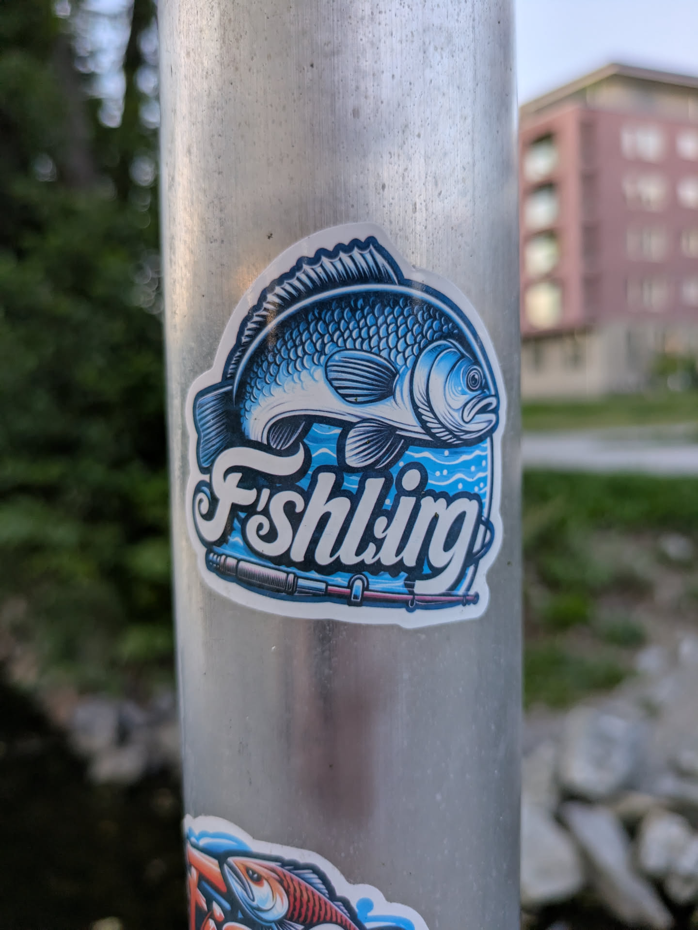 The sticker features a prominent, detailed blue and white fish, resembling a carp or similar freshwater species, depicted above stylized blue waves. The fish has intricate scale patterns and fins, with darker blue shading and white highlights. Below the fish, the word "Fishling" is written in a flowing, white script with a dark blue outline, giving it a dynamic appearance. A fishing rod, also in shades of blue and white, is positioned beneath the text. The entire design is enclosed within a thick, clean white border, making it stand out against the metallic pole it is attached to. The primary colors are various shades of blue, white, and black for outlines and details.