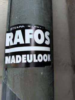 Street sticker The sticker is predominantly black with white text. The top line of text reads 'RAFOS' in large, bold, sans-serif capital letters with a slight blue or teal outline. Below this is a thin white horizontal line. The bottom line of text reads 'MADEULOOK' in similar bold, white, sans-serif capital letters. The sticker appears to be applied to a cylindrical, greenish-gray surface, possibly a pipe.