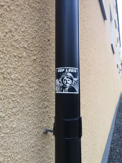 Street sticker A rectangular, high-contrast black and white sticker featuring a stylized depiction of an astronaut or cosmonaut. The central figure wears a space helmet, revealing a face with a prominent mustache and a determined expression. The background is a dark, starry sky with abstract white lines or rays emanating from the right side. At the top, a black banner contains the white text "» HOP LOUIE »". The overall aesthetic is graphic and bold, reminiscent of retro sci-fi or propaganda art.