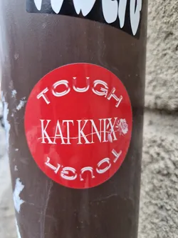 Red round sticker with white text. It says TOUGH KATKNIX TOUGH in a circular pattern. There is a small rose image next to the text KATKNIX. 