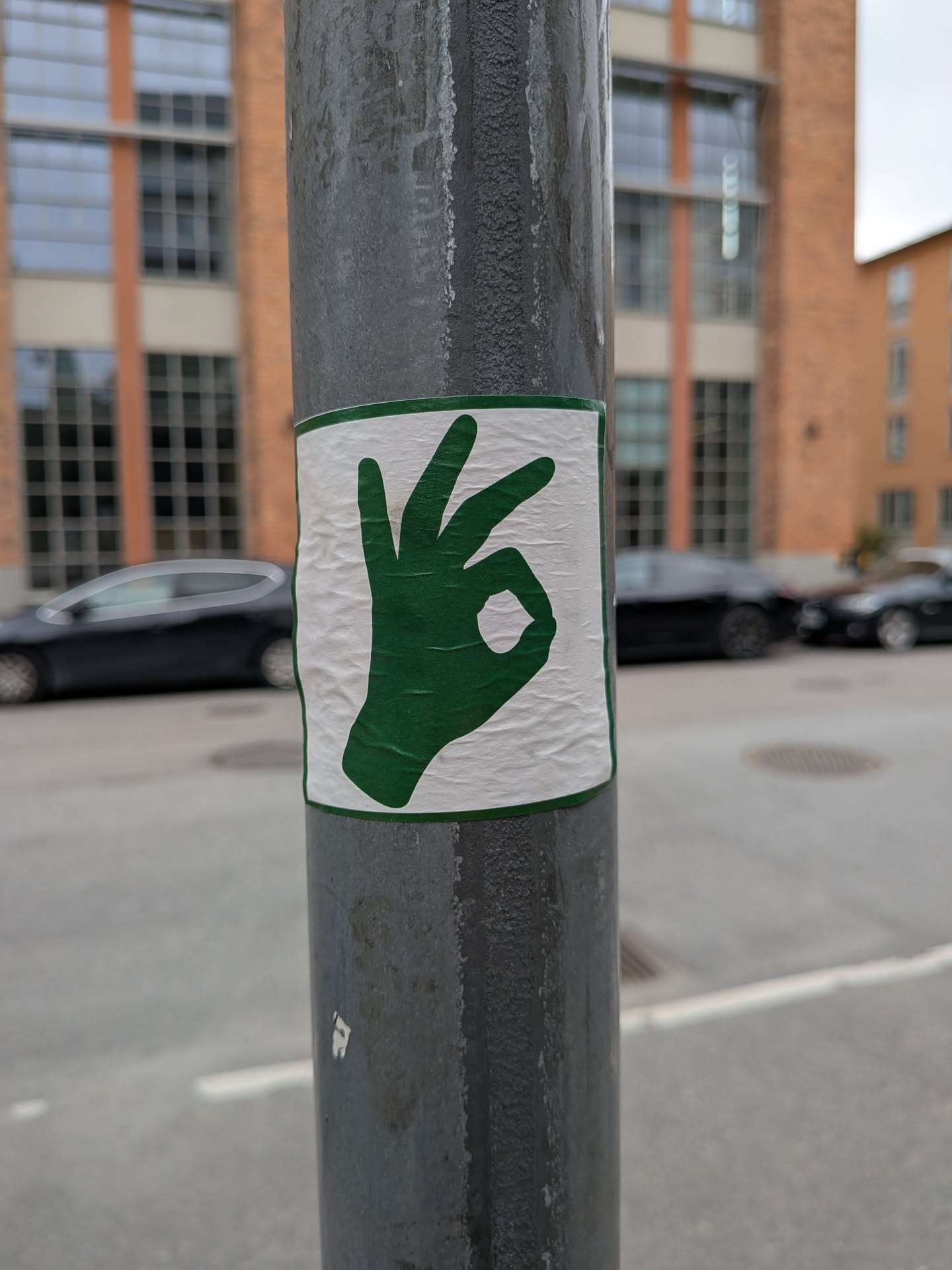 A square sticker with a thin green border and a white background, featuring a solid green silhouette of a hand making the 'OK' gesture. The sticker is affixed to a grey metal pole outdoors, showing some wrinkles and texture.