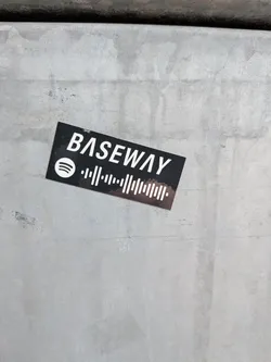 Street sticker A rectangular black sticker featuring white text and graphics. The word 'BASEWAY' is prominently displayed in a sans-serif font. To its left, the iconic white Spotify logo (three curved lines within a circle) is visible. Below 'BASEWAY' and extending to the right of the Spotify logo is a white Spotify code, which is a unique audio waveform pattern designed for scanning within the Spotify app. The sticker shows minor wear and tear, suggesting it's adhered to a textured, light gray surface.