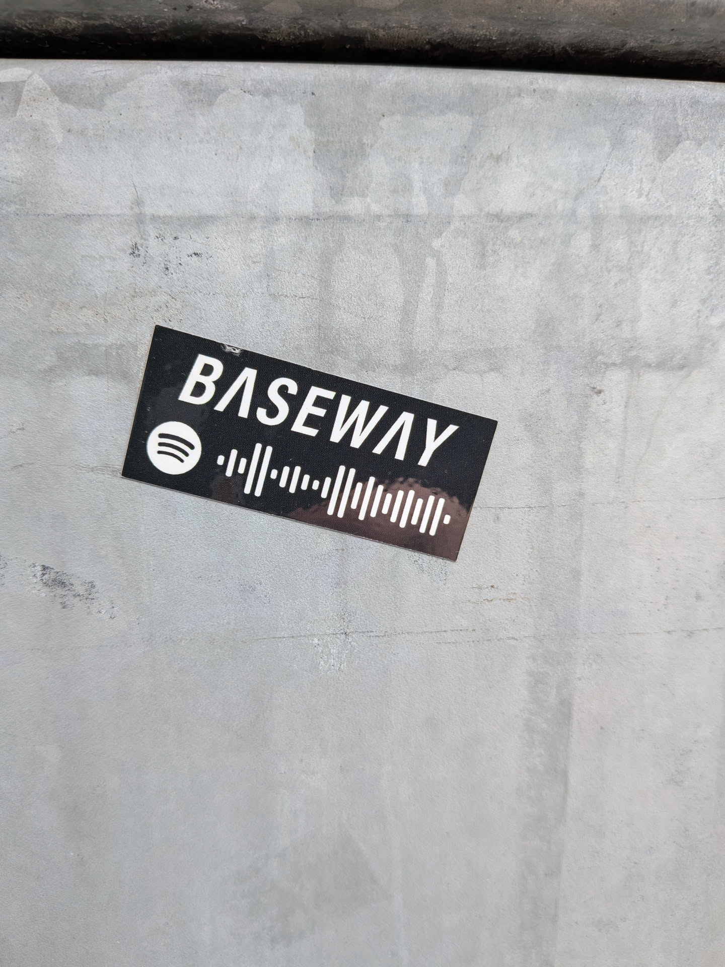 Street sticker A rectangular black sticker featuring white text and graphics. The word 'BASEWAY' is prominently displayed in a sans-serif font. To its left, the iconic white Spotify logo (three curved lines within a circle) is visible. Below 'BASEWAY' and extending to the right of the Spotify logo is a white Spotify code, which is a unique audio waveform pattern designed for scanning within the Spotify app. The sticker shows minor wear and tear, suggesting it's adhered to a textured, light gray surface.