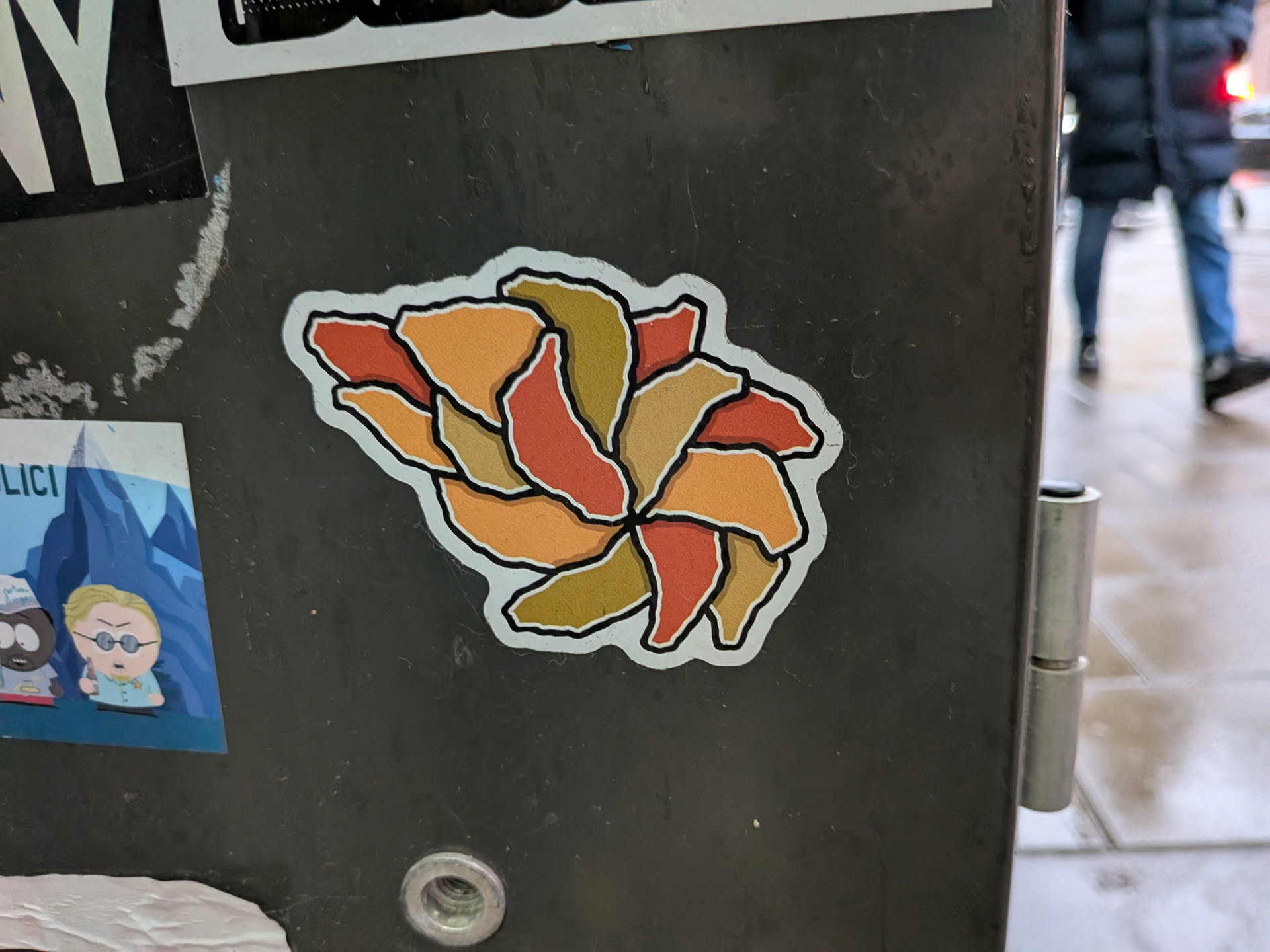 Street sticker The sticker is centrally located and features an abstract representation of a flower. It is composed of overlapping, petal-like shapes arranged in a circular pattern. The colors used are warm tones including shades of orange, red, and a muted olive green, with a distinct black outline around each shape and the overall sticker. The background of the sticker appears to be white, with a thin white border surrounding the entire design.