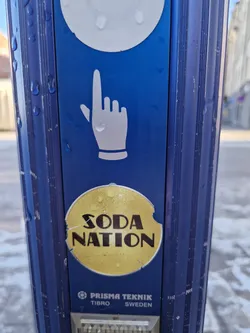 A round yellow sticker with the text "SODA NATION" in a stylized font. The sticker is slightly damaged with a small bite taken out of it. It is affixed to a blue public information pole. 