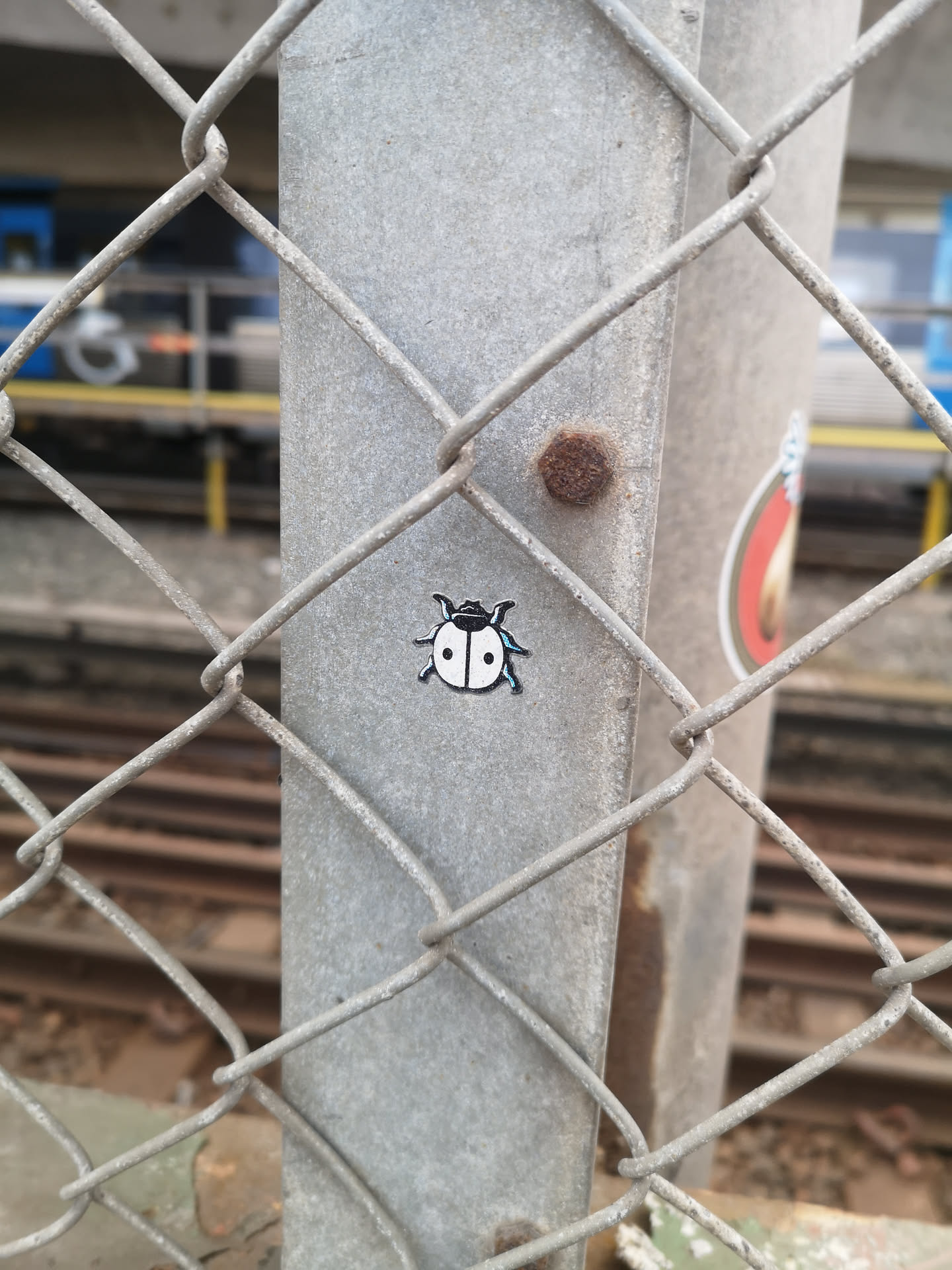The sticker features a minimalist, stylized depiction of a ladybug. Its body is predominantly white with two prominent black spots on its back. The head is black, and it has two small white eyes. Six thin black legs extend from its sides, and two small antennae protrude from its head. A subtle light blue outline or highlight is visible around the edges of its body, legs, and antennae, giving it a slightly glowing or digital appearance against the dark black outlines.