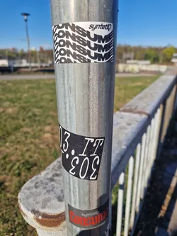 Street sticker The central focus of the image is a weathered, metallic pole adorned with three distinct stickers. The uppermost sticker is a white rectangle featuring the brand name "syntetiq" in a clean, black sans-serif font at its top. Below this, the word "CONSUME" is dynamically repeated four times in bold, black capital letters, arranged in a stacked, wavy pattern across the white background.
Mid-pole, a rectangular black sticker displays white text. The characters are "IT" followed by a period, and then what appears to be an inverted "303", visually transcribed as "E0E". The white text has a slightly worn or textured appearance against the matte black background.
The lowest visible sticker is partially obscured and features a dark, likely black or very dark grey, background. On this, the word "syntetiq" is visible in a smaller, white font, positioned above the word "Consume" which is rendered in bold, distressed red capital letters. The overall aesthetic of the stickers suggests an urban, graphic design style.