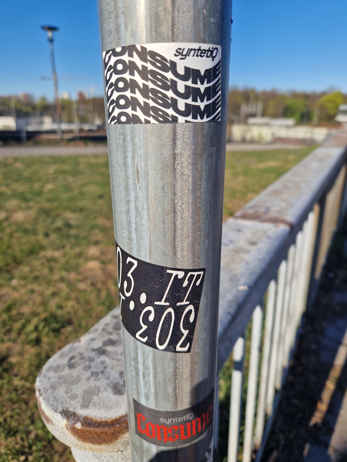 The central focus of the image is a weathered, metallic pole adorned with three distinct stickers. The uppermost sticker is a white rectangle featuring the brand name "syntetiq" in a clean, black sans-serif font at its top. Below this, the word "CONSUME" is dynamically repeated four times in bold, black capital letters, arranged in a stacked, wavy pattern across the white background.
Mid-pole, a rectangular black sticker displays white text. The characters are "IT" followed by a period, and then what appears to be an inverted "303", visually transcribed as "E0E". The white text has a slightly worn or textured appearance against the matte black background.
The lowest visible sticker is partially obscured and features a dark, likely black or very dark grey, background. On this, the word "syntetiq" is visible in a smaller, white font, positioned above the word "Consume" which is rendered in bold, distressed red capital letters. The overall aesthetic of the stickers suggests an urban, graphic design style.