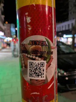 Street sticker A round sticker is affixed to a pole with reflective red and yellow stripes. The sticker features the name "ALYZZA" in a stylized font above an image of hands holding a fig, a lime and a green chili. The text "NEW MUSIC OUT NOW!" is printed on the sticker. There is also a QR code present.