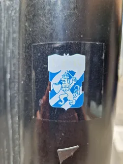 Street sticker A small, rectangular sticker featuring a blue and white coat of arms.  The design shows a crowned lion holding a heart, set against diagonal stripes.  The sticker appears to be affixed to a dark pole.