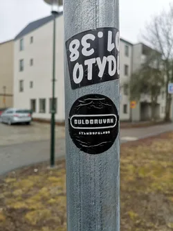 Street sticker The image displays two distinct monochrome stickers affixed to a textured, galvanized metal pole. The top sticker is rectangular, oriented diagonally, featuring a black background with bold white text. When rotated to be read correctly, the text appears to be a tag: 'OTYD' on the top line and '38.LO' on the bottom line. The bottom sticker is circular, also with a black background. It prominently features the word 'GULDGRUVAN' (Swedish for 'The Gold Mine') in white, stylized text enclosed within a white-outlined horizontal capsule shape. Below this, in smaller white text, is 'STANDSPELKLUBB' (Swedish for 'Tabletop Gaming Club'). Both stickers show minor creases and reflections, indicating they are physical objects adhered to the pole.