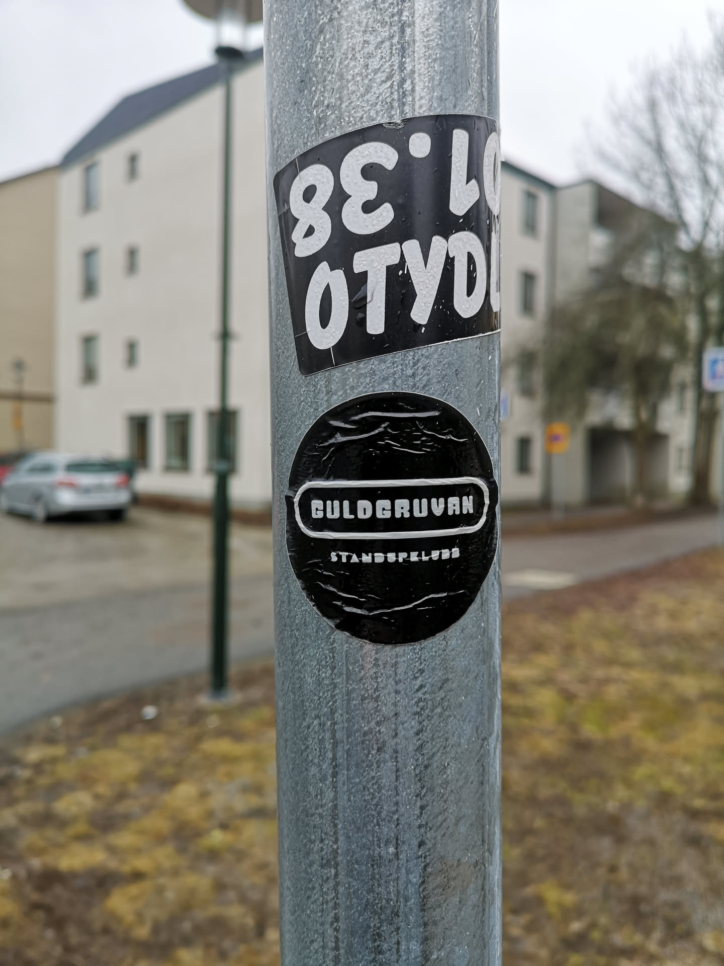 The image displays two distinct monochrome stickers affixed to a textured, galvanized metal pole. The top sticker is rectangular, oriented diagonally, featuring a black background with bold white text. When rotated to be read correctly, the text appears to be a tag: 'OTYD' on the top line and '38.LO' on the bottom line. The bottom sticker is circular, also with a black background. It prominently features the word 'GULDGRUVAN' (Swedish for 'The Gold Mine') in white, stylized text enclosed within a white-outlined horizontal capsule shape. Below this, in smaller white text, is 'STANDSPELKLUBB' (Swedish for 'Tabletop Gaming Club'). Both stickers show minor creases and reflections, indicating they are physical objects adhered to the pole.