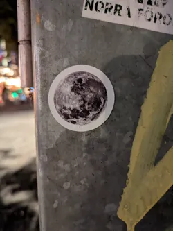 Street sticker The sticker is circular and features a detailed, monochromatic image of the moon. The lunar surface, with its craters and maria, is clearly visible, rendered in shades of gray and black against a white border. The sticker is affixed to a rough, metallic surface, possibly a pole, which also has some yellow paint markings.
