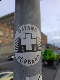 Street sticker A circular sticker with a black and white design adhered to a metal pole. The outer edge of the circle is a thick black line. Inside, the text 'GATANS' arcs over the top, and 'FÖRBUND' arcs under the bottom, both in black block letters. The center of the sticker features a stylized white cross, which appears to be overlaid on a graphic representation of buildings. These buildings, depicted with dark lines suggesting windows and structure, flank the cross on either side. The sticker has a slightly worn, textured appearance, indicating it is likely a real sticker that has been exposed to the elements.
