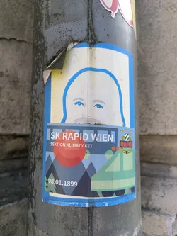 A slightly peeling sticker affixed to a pole. The sticker has a blue border and depicts a portrait at the top, and an illustration of a landscape with geometric mountains and a train-like object at the bottom. The sticker is labeled "SK RAPID WIEN" and "SEKTION KLIMATICKET". There is text reading "08.01.1899" at the bottom left.