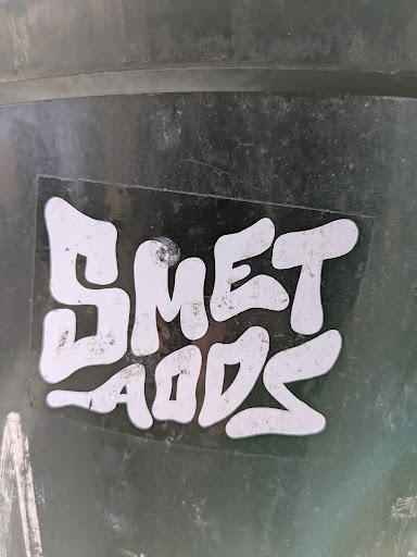 Street sticker A close-up of a sticker with the text "SMET LADS" in a stylized, rounded font. The sticker is black with white lettering, affixed to a dark, possibly metallic or plastic, surface.