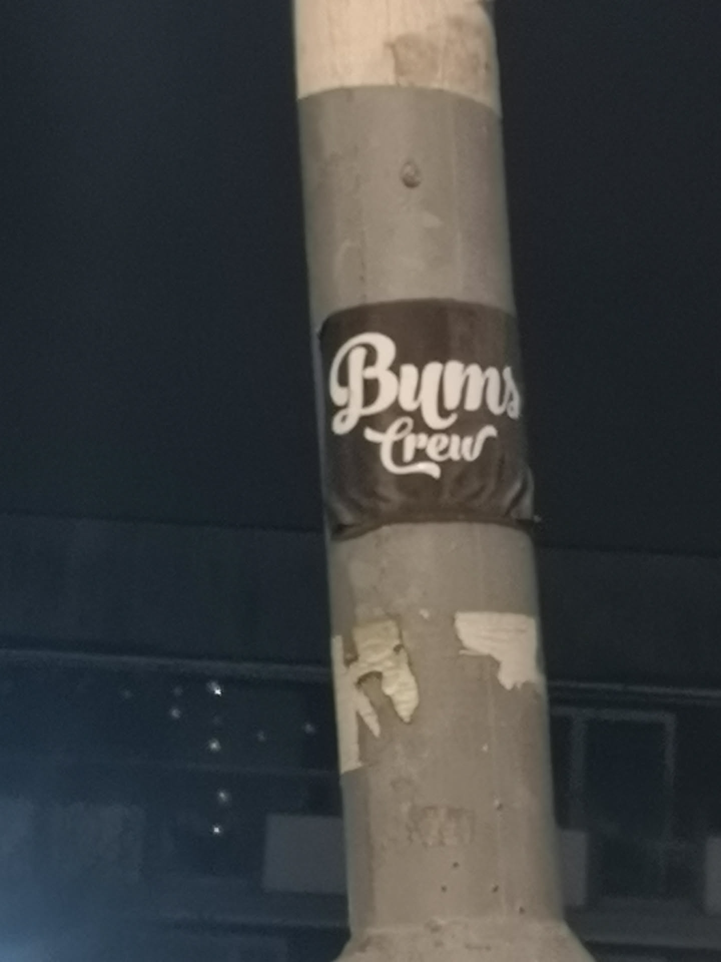 A rectangular sticker with a dark brown background, applied to a light grey pole. The sticker features the text 'Bums Crew' in a flowing, white script font. 'Bums' is larger and positioned above 'Crew', which is slightly smaller and centered below it. The sticker shows some minor wrinkles and wear, consistent with being affixed to an uneven surface.