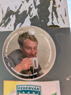 Street sticker This is a round sticker on a wall. It features a man drinking from a mug with the word "GAUGAU" on it. The sticker has text around the edge, including "WER HOD DENN GEBURTSTOG?", "SCHURZENJAGER & WURMBOODER" and "GAUGAU REINHEITSGEBOT EST. 1996".