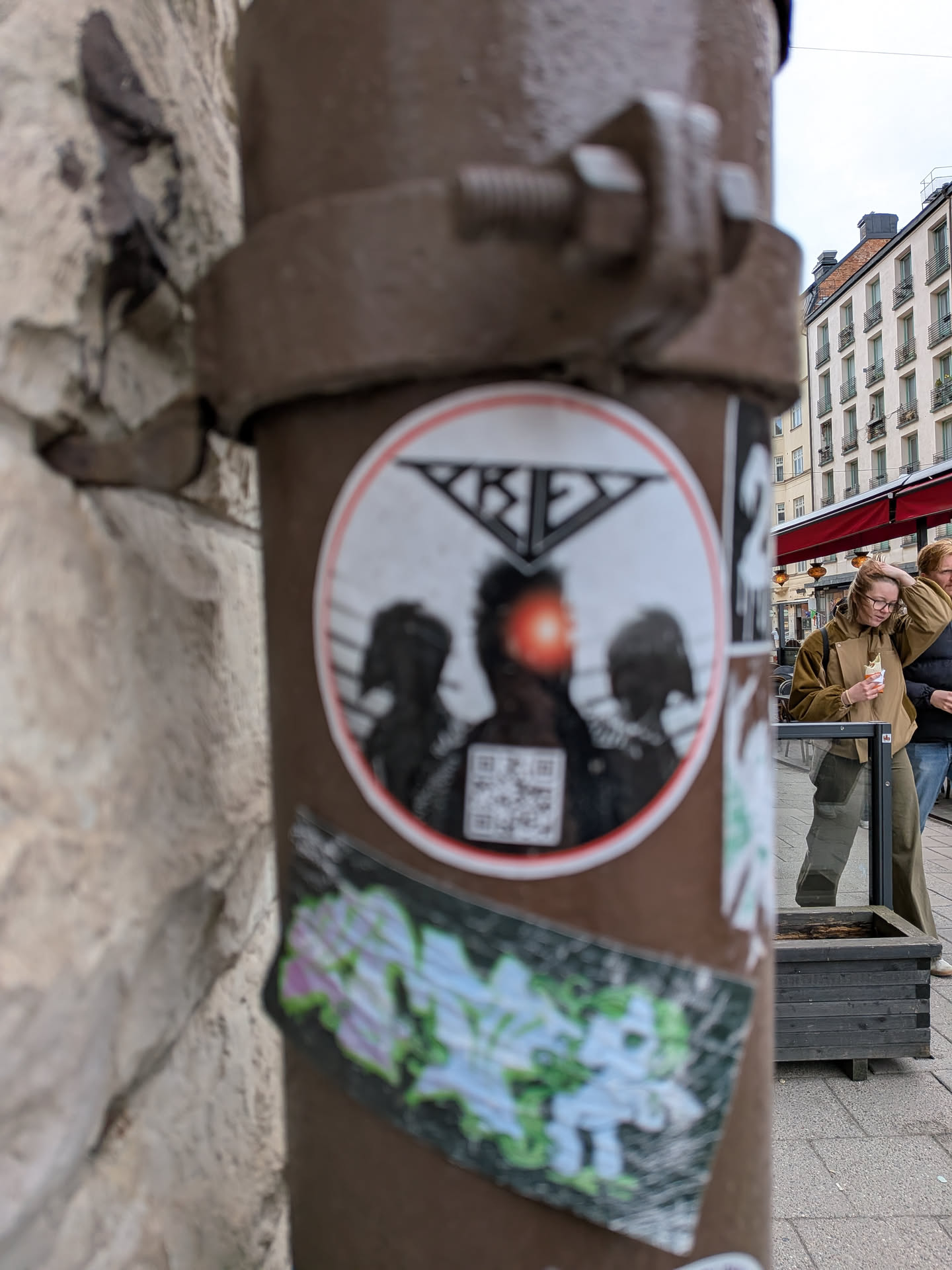 The sticker is circular with a white background and a prominent red outer border. It depicts three dark, silhouetted figures, with the central figure being slightly larger and having a bright orange-red glow emanating from its head or face area. Above the figures, there is a black, stylized geometric text that appears to spell 'KREYZ' or a similar word, forming a triangular shape. Below the central figure, a black and white QR code is visible. The overall aesthetic is somewhat edgy and urban, adhered to a brown metal pipe.