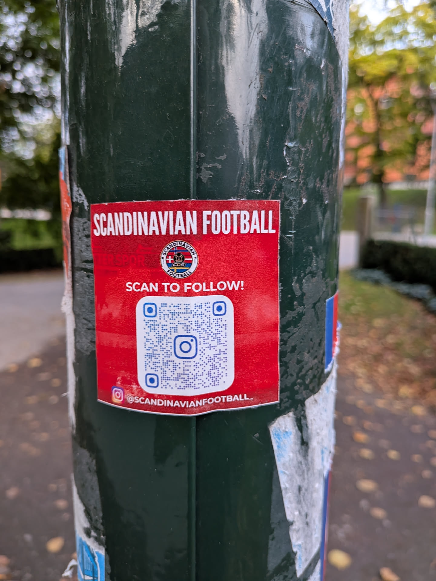 A rectangular red sticker stuck to a dark green pole. It features the text 'SCANDINAVIAN FOOTBALL' at the top in white, a circular logo containing Nordic flags, the call to action 'SCAN TO FOLLOW!', a large QR code with an Instagram logo in the center, and the Instagram handle '@SCANDINAVIANFOOTBALL' at the bottom.