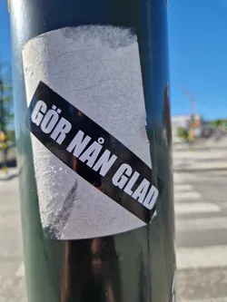 Street sticker A sticker with the text "GÖR NÅN GLAD" which translates from Swedish to "Make someone happy".  The sticker is attached to a dark green pole, likely outdoors.
