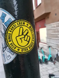 A round yellow sticker with a peace sign and the text "Otek Bingo Kaksi Crew" repeating around the edge. The sticker is applied to a dark pole outdoors, with some other stickers visible nearby.  There are also several scooters visible in the background.