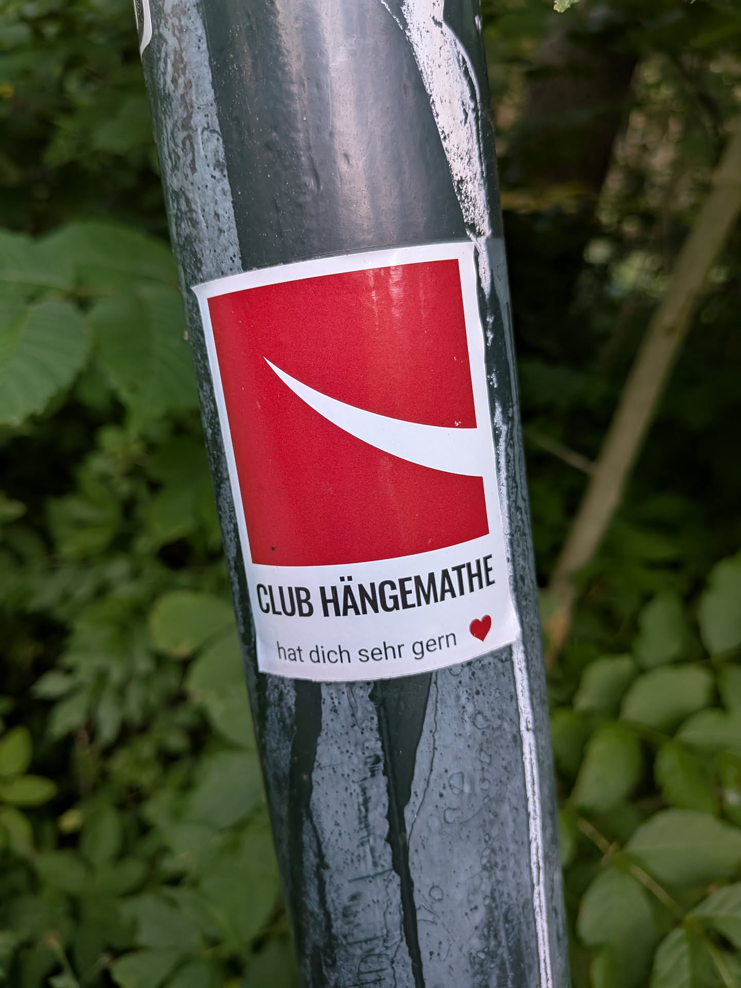 A rectangular sticker on a pole. It features a red square with a white curved swoosh representing a hammock. Below, black text reads 'CLUB HÄNGEMATHE' and 'hat dich sehr gern' with a small red heart.