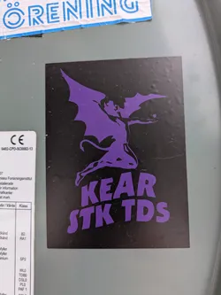 Street sticker A rectangular sticker with a black background featuring a prominent purple graphic. The graphic depicts a stylized, winged demonic figure with a tail, striking a pose similar to album art from the band Black Sabbath (specifically, the 'Master of Reality' aesthetic). The figure is holding a small five-pointed star in its outstretched hand. Below the figure, in bold, slightly wavy purple letters, are the words 'KEAR' on the first line and 'STK TDS' on the second line. The sticker is applied to a grey, flat surface.