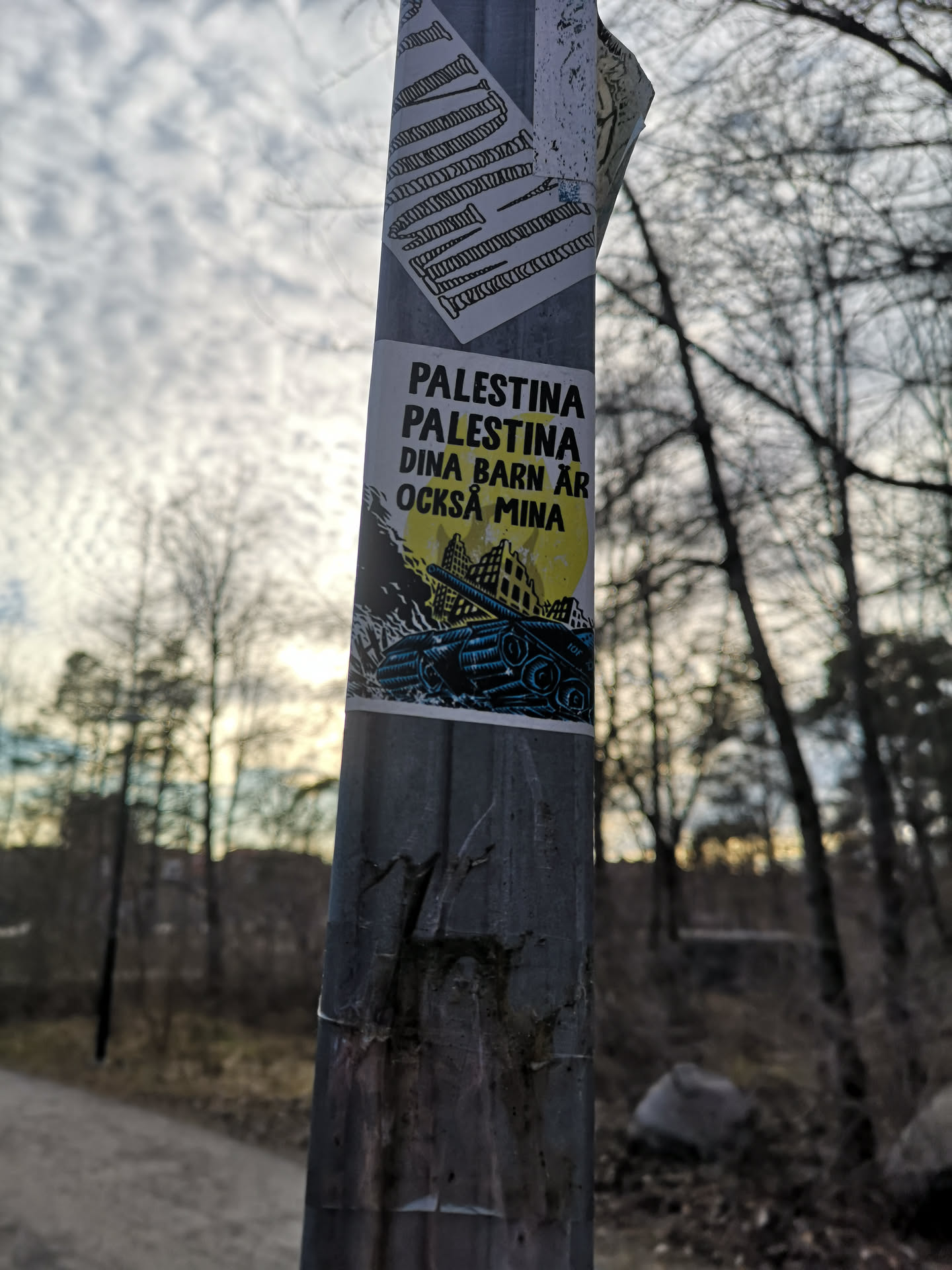 The sticker is a rectangular piece adhered to a metal pole. It features a stark graphic design with a primary color palette of black, yellow, and blue. The upper portion displays bold, black uppercase text on a white background. Below the text, a large, bright yellow circle, possibly representing a sun or moon, dominates the background. Overlaid on this yellow circle and extending downwards is a dark, silhouetted landscape of what appears to be destroyed or war-torn buildings. In the foreground, a detailed illustration of a blue military tank is prominently featured, moving across the lower part of the scene. The overall aesthetic is impactful and conveys a strong political or social message.