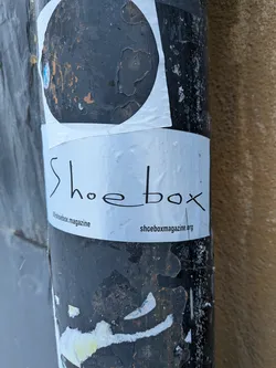 A rectangular white sticker featuring the word 'Shoebox' in a stylized, handwritten-like black font, centered horizontally. Below this, in smaller black text, '@shoebox.magazine' is on the left and 'shoeboxmagazine.org' is on the right. The sticker is slightly wrinkled and shows minor wear, adhered to a heavily weathered, dark-colored pole with peeling paint. Above this sticker, a larger, partially visible white sticker with a black circular design is also present.