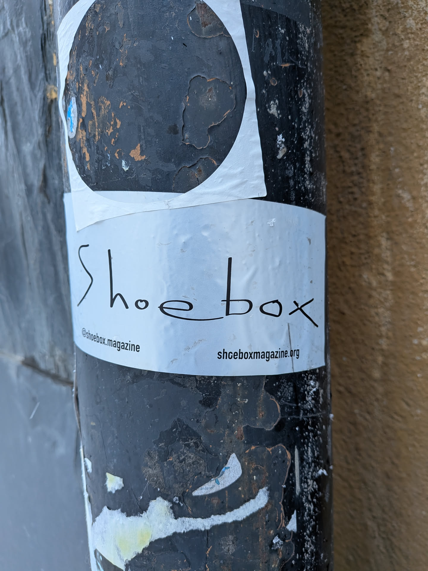 Street sticker A rectangular white sticker featuring the word 'Shoebox' in a stylized, handwritten-like black font, centered horizontally. Below this, in smaller black text, '@shoebox.magazine' is on the left and 'shoeboxmagazine.org' is on the right. The sticker is slightly wrinkled and shows minor wear, adhered to a heavily weathered, dark-colored pole with peeling paint. Above this sticker, a larger, partially visible white sticker with a black circular design is also present.