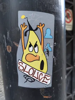 A sticker featuring a cartoon illustration of a yellow pear-like character with arms raised in anger. It has a black hat, orange arms, and a grumpy facial expression. The word 'SLOUCE' is written in stylized lettering on a banner below the character. Two small, light blue figures are positioned on the sticker near the character, appearing to observe. The sticker is affixed to a dark gray pole.