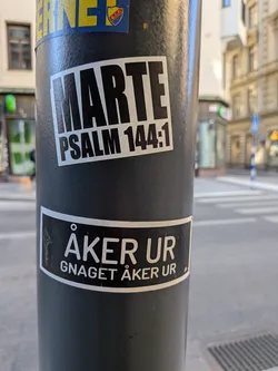 The sticker is rectangular with slightly rounded corners, featuring a stark black-on-white design. The word 'MARTE' is prominently displayed in large, bold, sans-serif capital letters across the top. Below it, in a smaller but still bold sans-serif font, is the text 'PSALM 144:1'. The sticker appears to be made of vinyl, showing minor signs of wear and adherence to the textured surface of the pole.